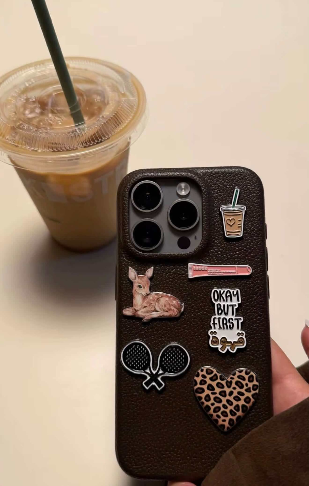 Six glossy metal stickers layered on a black leather phone case: an iced coffee, a fawn, crossed tennis rackets, a lip gloss, a lettering sticker and a leopard print heart